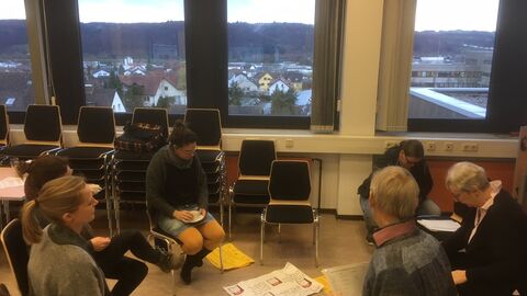 Teilnehmer und Teilnehmerinnen sitzen in kleine Gruppen zusammen und bearbeiten Aufgaben, die auf Plakate geschrieben sind.