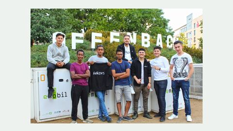 Zu stehen sind acht junge Männer die vor einer Mauer stehen. Hinter ihnen sieht man grüne Pflanzen und das Wort Offenbach aus großen weißen Buchstaben. Einer der Jungen hält einen schwarzen Pulli mit der Aufschrift HeRoes in der Hand.