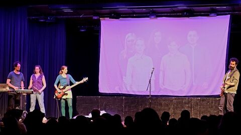Szene aus dem Theaterstück Musikalische Szene aus dem Theaterstück. Auf der Bühne befinden sich Schauspielerinnen und Schauspieler mit Musikinstrumenten. Im Hintergrund sind auf einer Leinwand Kinder zu sehen.