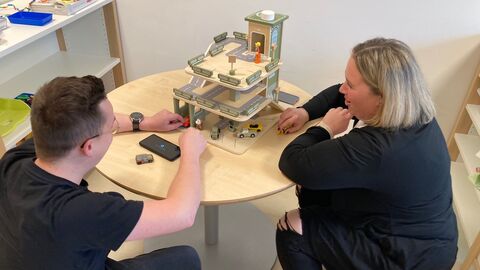 Workshopteilnehmer bei der Ausführung einer Praxisaufgabe Personen hocken vor einen niedrigen Tisch auf dem ein Spielzeugparkhaus mit Spielzeugautos und -figuren stehtsteht.