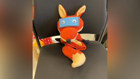 Die Sympathiefigur MEKOKI, eine Figur mit dem Aussehen eines Fuchses mit einer Brille in Form eines Smartphones, sitzt auf einem Stuhl