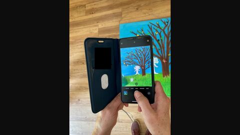 Mittels Smartphone wird ein Gemälde mit grüner Wiese und Baum fotografiert. Links neben dem Baum steht eine selbstgezeichnete Figur. 