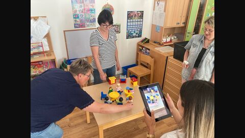 Erziehrinnen stehen um einen Tisch mit Spielzeugfiguren und Spielzeugmöbel. Mittels Tablet hält eine Erzieherin die Situation bildlich fest.