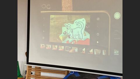 Auf einer Leinwand wird das Display eines Smartphones gezeigt. Auf dem Display ist ein gemalter Hund und eine Schokoriegel zu sehen. 