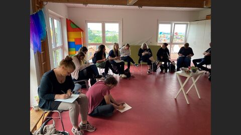 Mehrere Personen sitzen in einem Raum, teilweise auf Stühlen, teilweise auf dem Fußboden und malen.