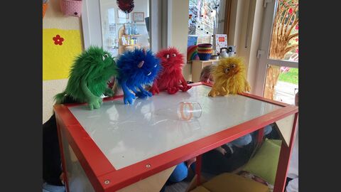 Auf einem Tisch sitzen vier Handpuppen, Monster, vor einem umgefallenen Glas. Auf dem Tisch ist eine Wasserpfütze. Hinter dem Tisch sind einzelne Körperteile von personen zu erkennen, welche die Handpuppen spielen.