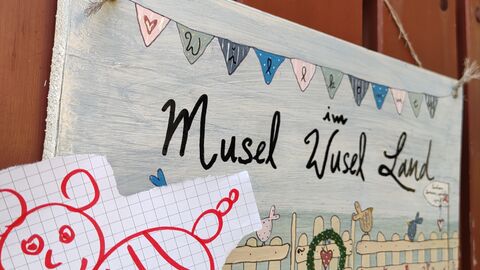 Ein selbst gemaltes Schild mit dem Schriftzug Musel-Wusel-Land hängt an einem Zaun.