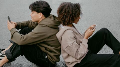 zwei Personen sitzen mit angewinkelten Beinen Rücken an Rücken auf dem Boden. Jede Person hält ein Smartphone in der Hand.